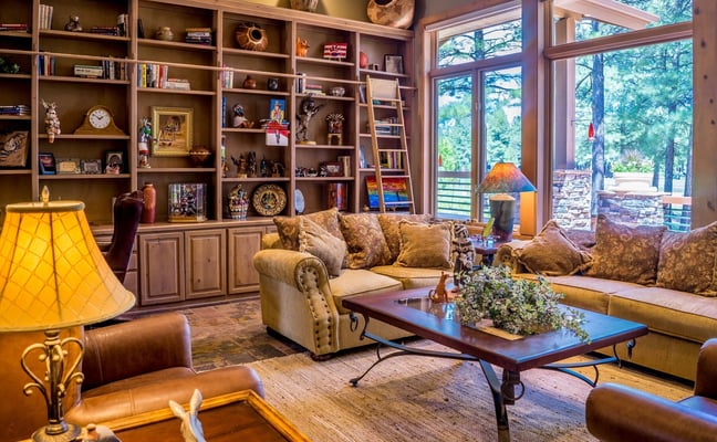 Luxury living room with built-in shelving, upholstered seating, dark wood coffee table, and large windows overlooking wooded landscape.
